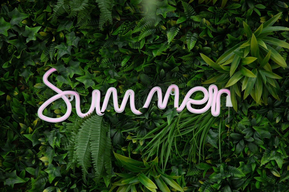 A pink neon sign reading Summer mounted on a wall of artificial green foliage