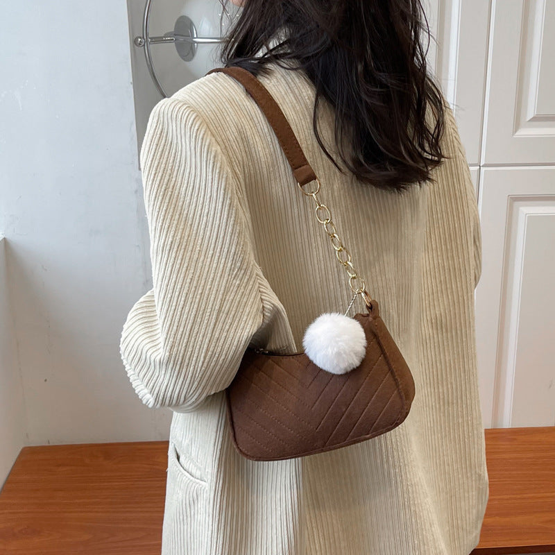 Braune Wildleder-Handtasche mit Zickzack-Muster, weißem Bommel und goldener Kette, getragen von einer Frau mit beige Cordjacke. IsabellBoutique030